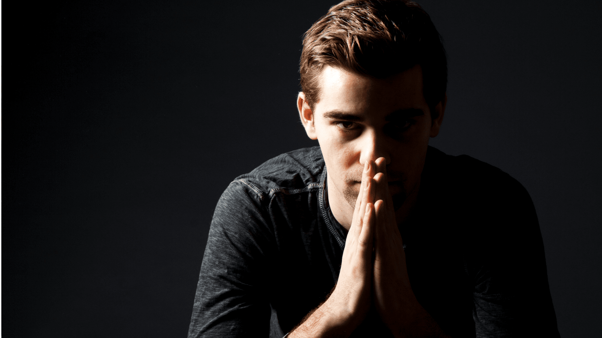 Man in shadowed light with focused gaze and clasped hands, conveying emotional intensity